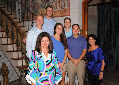 A family of seven people smiling for a photo in a living room with a staircase and tapestry in the background.