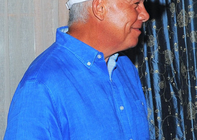 Man in a blue shirt and white cap holding a wine glass, smiling and turning away from the camera with curtains in the background.