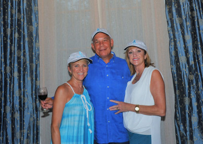 Three adults posing in front of a curtain, two women wearing white hats and a man in a blue shirt, holding a glass of wine.