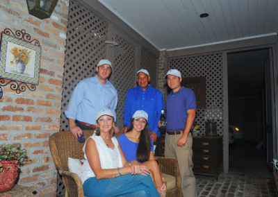 Five adults smiling for a photo on a porch at night, three men standing, two women seated, all wearing festive hats.