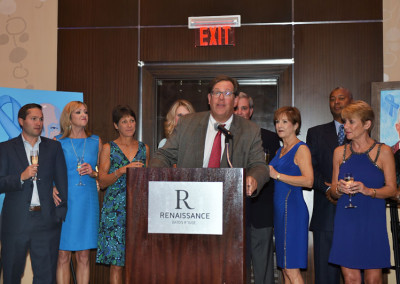 A man speaks at a podium during an event at renaissance baton rouge, with a group of people standing behind him.