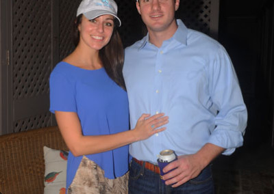 A couple stands together on a patio at night, the man holding a beverage can, both wearing baseball caps.