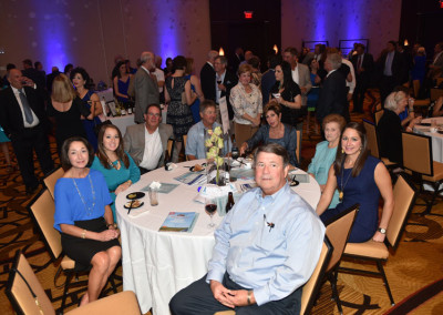 Group of people at a gala event, sitting and standing around tables in a banquet hall with blue lighting.