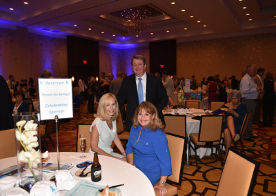 Three people smiling at a table during a formal event in a ballroom with attendees and a 'reserved' sign indicating sponsorship.