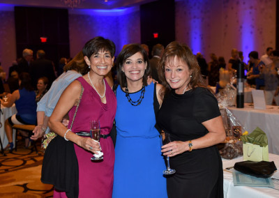 Three women smiling at a gala event, holding wine glasses, with a blurred background of guests and blue lights.