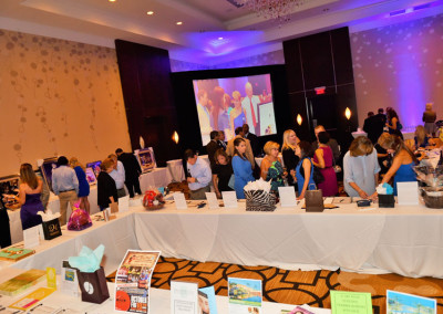 People browsing items at a charity auction event in a hotel ballroom with a large screen displaying a live auction.