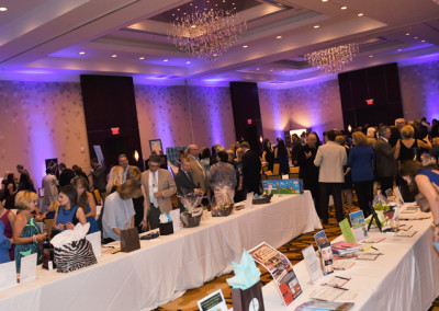 People mingling at a charity auction event in a hotel ballroom with items displayed on tables.