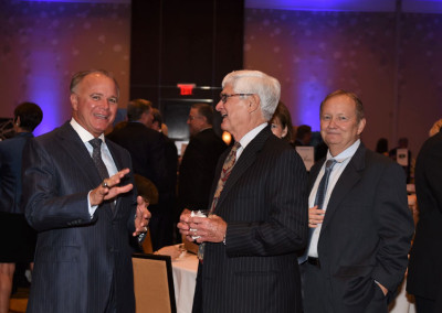 Three senior businessmen in suits conversing and laughing at an indoor corporate event with blue lighting in the background.
