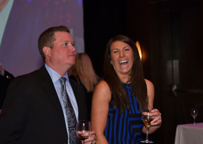 Two people laughing at an event, the woman holding a wine glass and the man in a suit looking to the side.