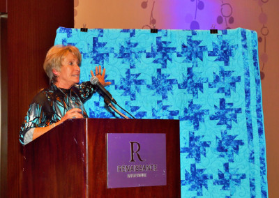 A woman speaks at a podium with a microphone, against a backdrop featuring a blue jigsaw puzzle design, at a renaissance hotel event.