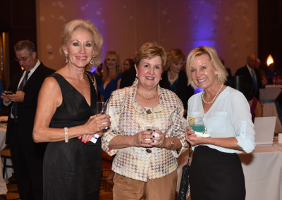 Three women holding drinks and smiling at a social event with other guests and purple lighting in the background.