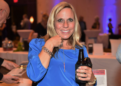 Woman in blue blouse smiling at a social event, holding a beer bottle, with indistinct guests and blue lighting in the background.