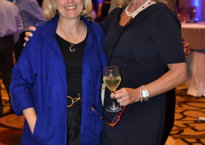 Two women smiling at a formal event, one holding a glass of wine. one wears a black dress and the other a blue jacket.