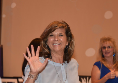 A smiling woman waving her hand, standing at a podium in a conference hall, with another woman visible in the background.