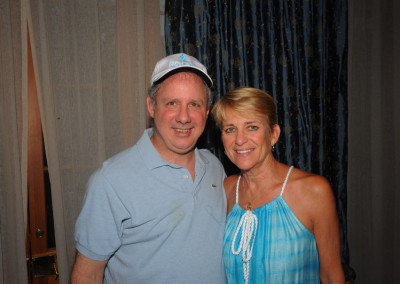 A man in a cap and a woman in a blue dress smiling for a photo inside a room with curtain backdrop.
