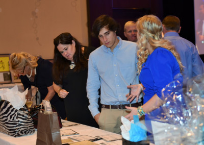 Three people examining items on a table at a charity event, with other attendees and decorations in the background.