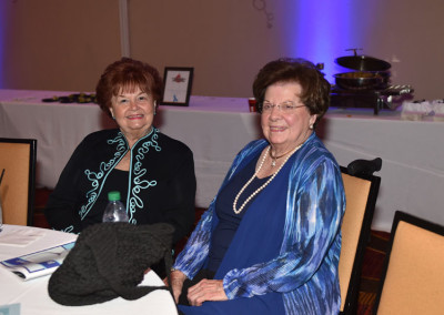 Two elderly women sitting at a banquet table, smiling at the camera, with a softly lit background featuring a buffet station.
