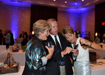 Two women and a man in formal attire viewing a smartphone together at an indoor event, with a decorated table in the background.
