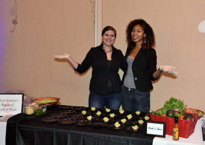 Two women standing behind a food stall, presenting guacamole dishes at an event.