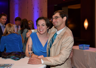 A smiling man and woman in formal attire sitting together at a social event, with people and purple lighting in the background.
