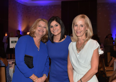 Three women smiling at a formal event, standing closely together in a room with dim lighting and a starry backdrop.
