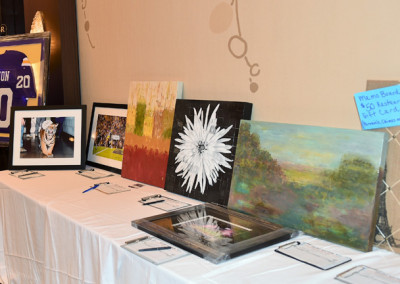 Silent auction display featuring paintings, a sports jersey, and a framed photo on a table with bid sheets.