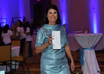 Woman in a sparkly blue dress holding a booklet, walking at an event with tables and blue lighting in the background.