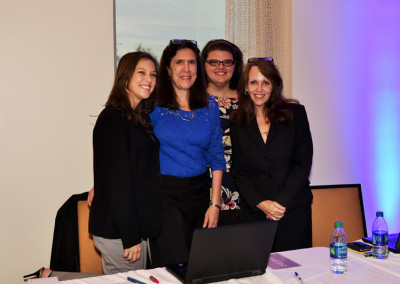 Four women smiling at a conference table lit by blue ambient light, with a laptop and water bottles in front of them.