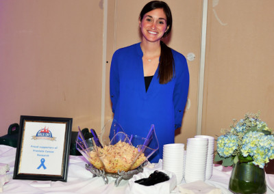 A smiling woman stands behind a table with snacks and a prostate cancer awareness sign at an event.