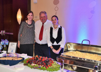 Three people posing together at a catering event with a food table featuring fruit and warm dishes.