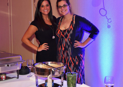 Two women smiling at a social event, standing beside a buffet table with food warmers and a wine glass in the foreground.