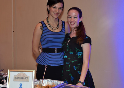 Two women smiling at a charity event stand in front of a food table with a sign supporting prostate cancer research.