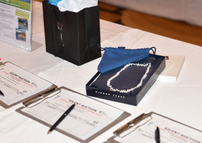 A silent auction table with registration forms, pens, and gift items including a pearl necklace and decorative bags.