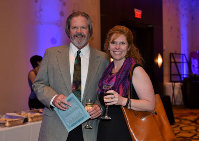 A man and a woman smiling at a formal event, the man holding a booklet and the woman holding a wine glass.