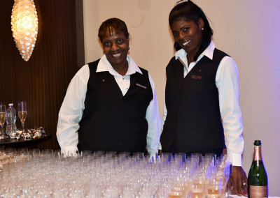 Two smiling female servers in uniform standing behind a table with rows of champagne glasses and a bottle.