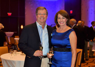 A man and a woman holding wine glasses at a formal event, smiling at the camera, with blue lighting in the background.