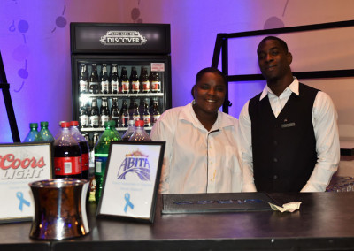 Two bartenders, a woman and a man, standing behind a bar with beverages and promotional signs.