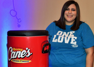 A woman in a blue "one love" t-shirt smiles beside a red raising cane's chicken fingers trash can against a purple wall.