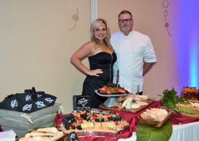 A woman and a chef standing behind a buffet table filled with an assortment of cheeses, fruits, and hors d'oeuvres at an event.