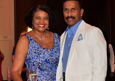 A man and a woman smiling at a formal event, with the woman in a blue dress and the man in a white suit jacket and blue tie.