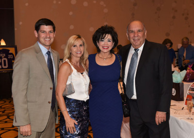 Four people smiling at a social event, two men in suits and two women in dresses, standing together in a room with a decorated backdrop.