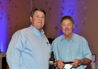 Two middle-aged men in blue shirts holding a beer and a cellphone, standing together and smiling at a social event.