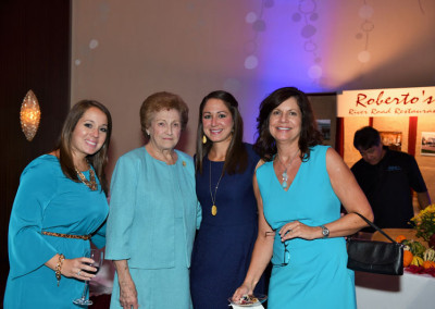 Four women in matching blue dresses holding drinks at a formal event, with a restaurant sign in the background.