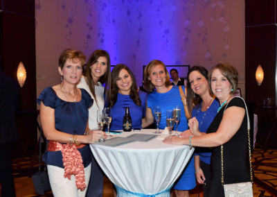 Six women smiling at a social event, gathered around a high table with wine glasses and a bottle.