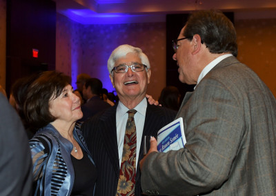 Two men and a woman engaging in conversation at a formal event, one man holding a book.