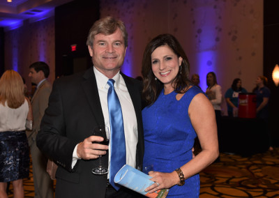 A man and a woman in formal attire posing at a social event, the man holding a glass of wine and the woman clutching a clutch purse.