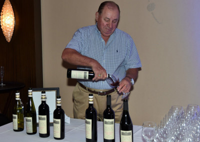 A man pours wine from a bottle into a glass at a wine tasting event, with multiple wine bottles and glasses arranged on a table.