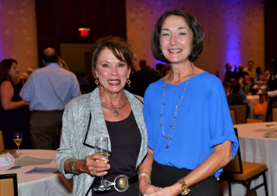 Two women smiling at a social event, one holding a glass of wine, both dressed in semi-formal attire.