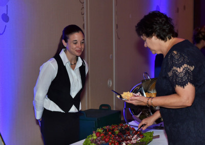 A waitress in a vest and tie observes a woman in a lacy black dress serving herself at a buffet.