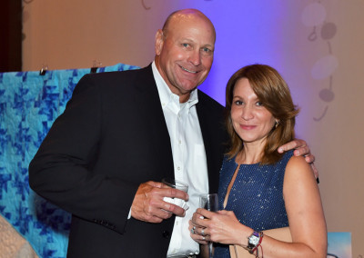 A man and a woman holding drinks, smiling at a formal event, with a decorative blue background.